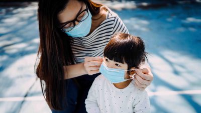 Young mother putting on surgical mask for little daughter in the playground to prevent the spread of cold and flu and viruses